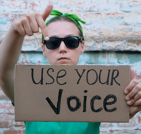 woman with dark glasses and green bandana and shirt pointing to a sign that say "use your voice"